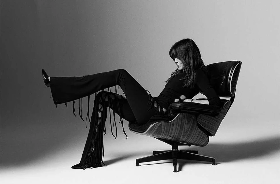 Monochrome portrait of a woman in a long-sleeved top and fringe pants reclining in a dark-wood chair against a gradient background.