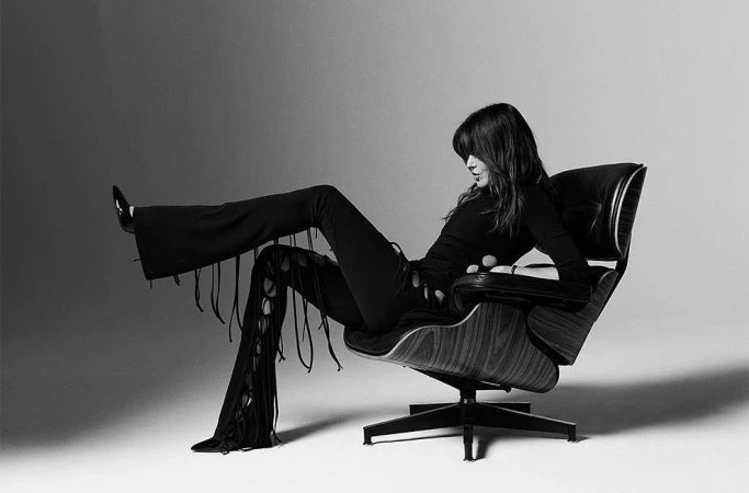 Monochrome portrait of a woman in a long-sleeved top and fringe pants reclining in a dark-wood chair against a gradient background.