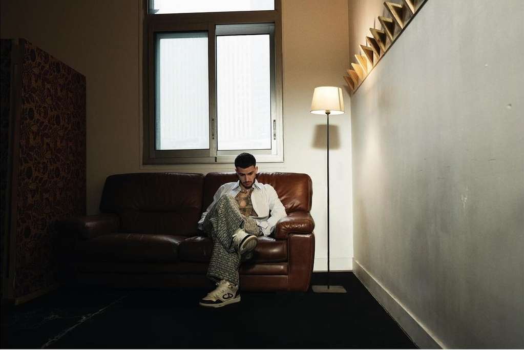 Stylish adult man sitting on a brown leather couch in a dimly lit room with a window, patterned wall, floor lamp, and wall decoration.