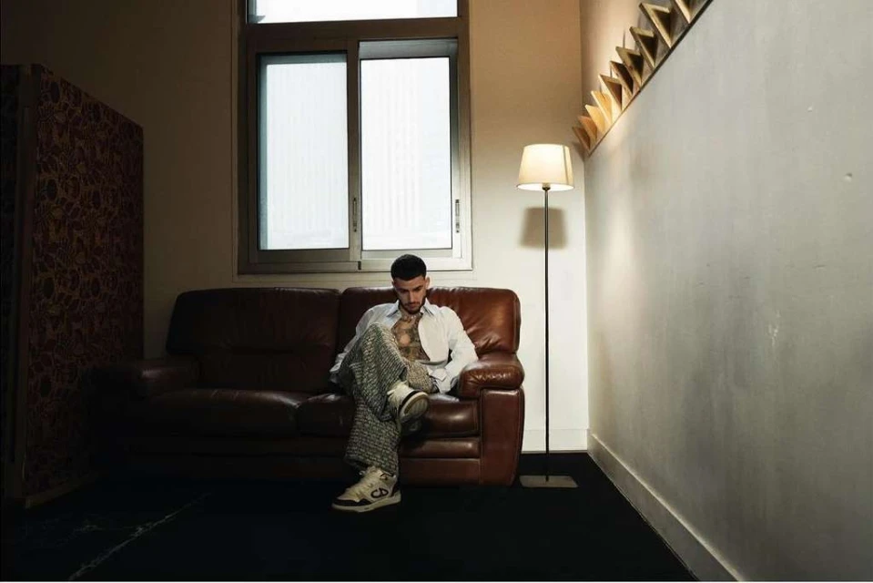 Stylish adult man sitting on a brown leather couch in a dimly lit room with a window, patterned wall, floor lamp, and wall decoration.