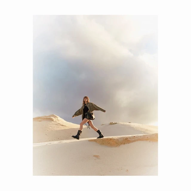 A woman with long brown hair strides across sand dunes in a fashionable outfit, featuring a jacket, shorts, and boots, set against a cloudy sky.