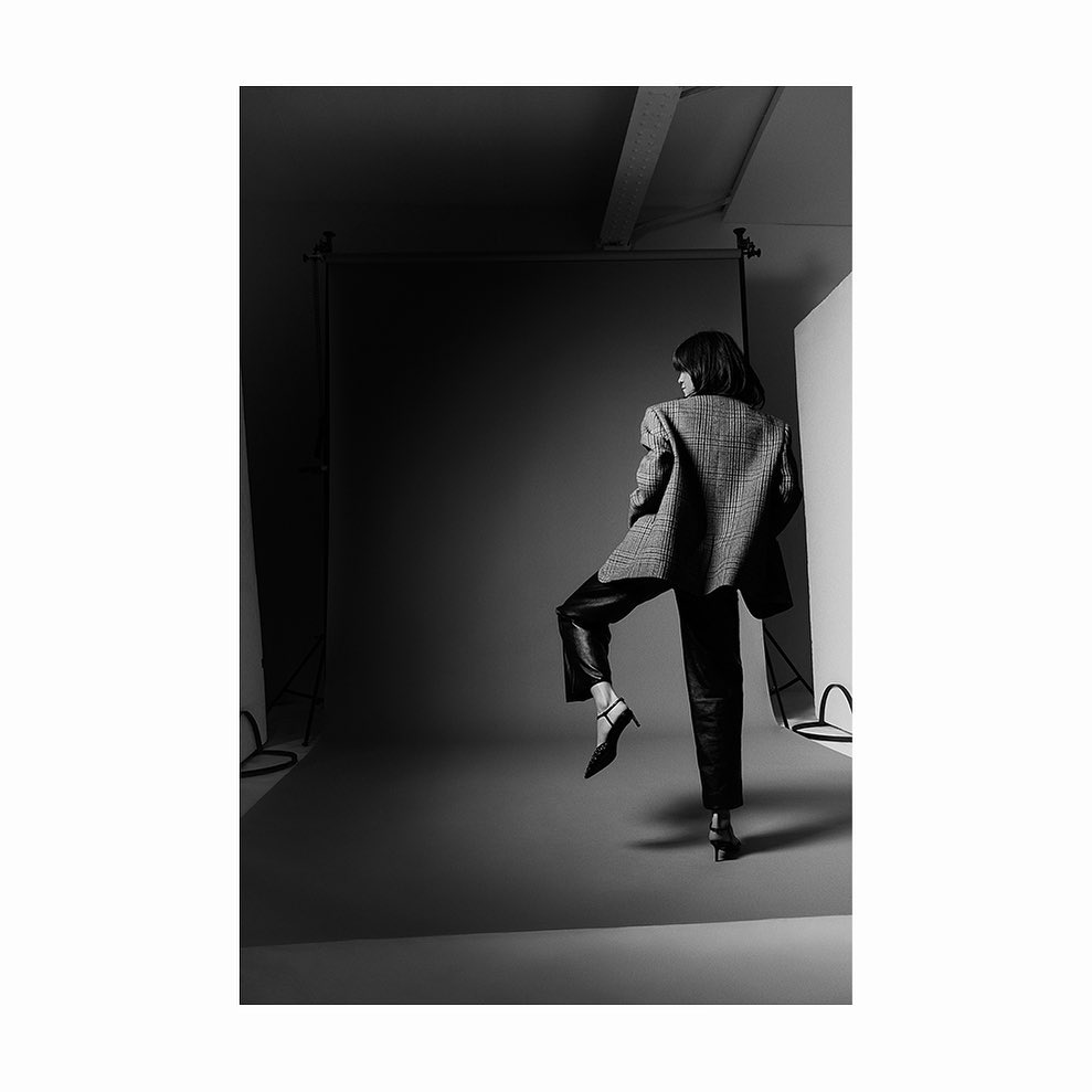 Woman in a houndstooth blazer and leather pants poses in a photo studio against a grey backdrop, highlighted by studio lighting.