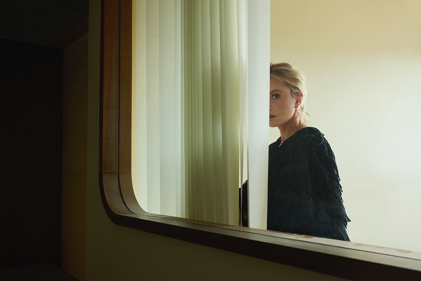 Elegant woman with blonde hair stands behind a window with vertical blinds, reflecting off a mirror in a sophisticated indoor setting.