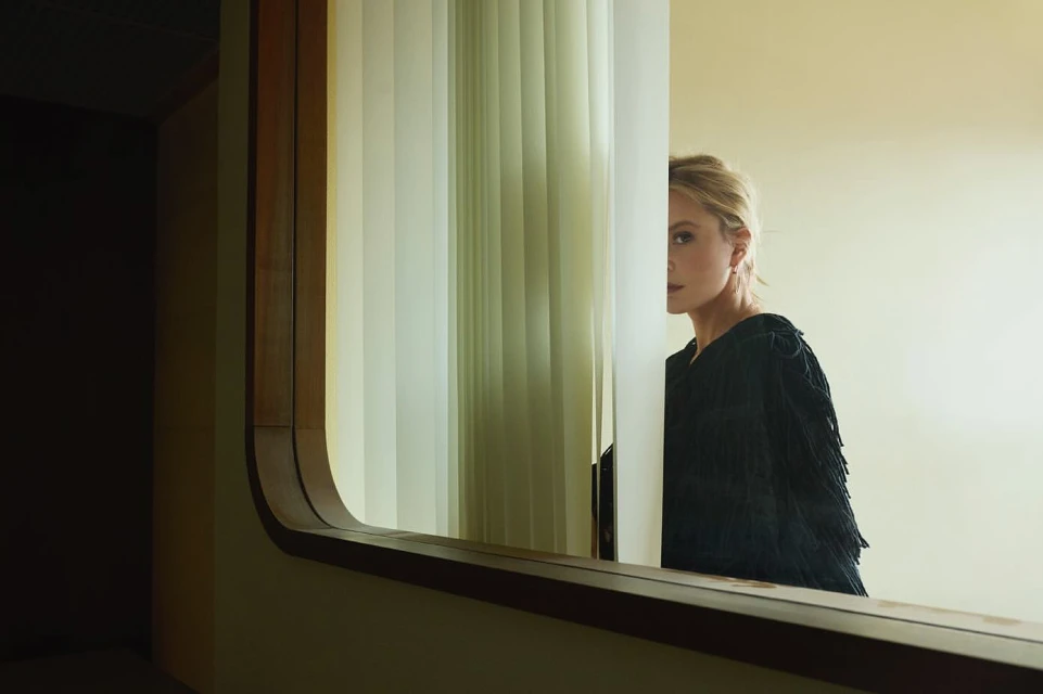 Elegant woman with blonde hair stands behind a window with vertical blinds, reflecting off a mirror in a sophisticated indoor setting.