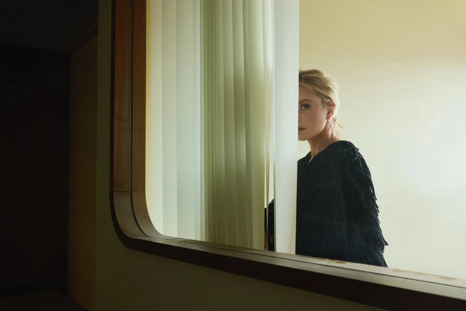 Elegant woman with blonde hair stands behind a window with vertical blinds, reflecting off a mirror in a sophisticated indoor setting.
