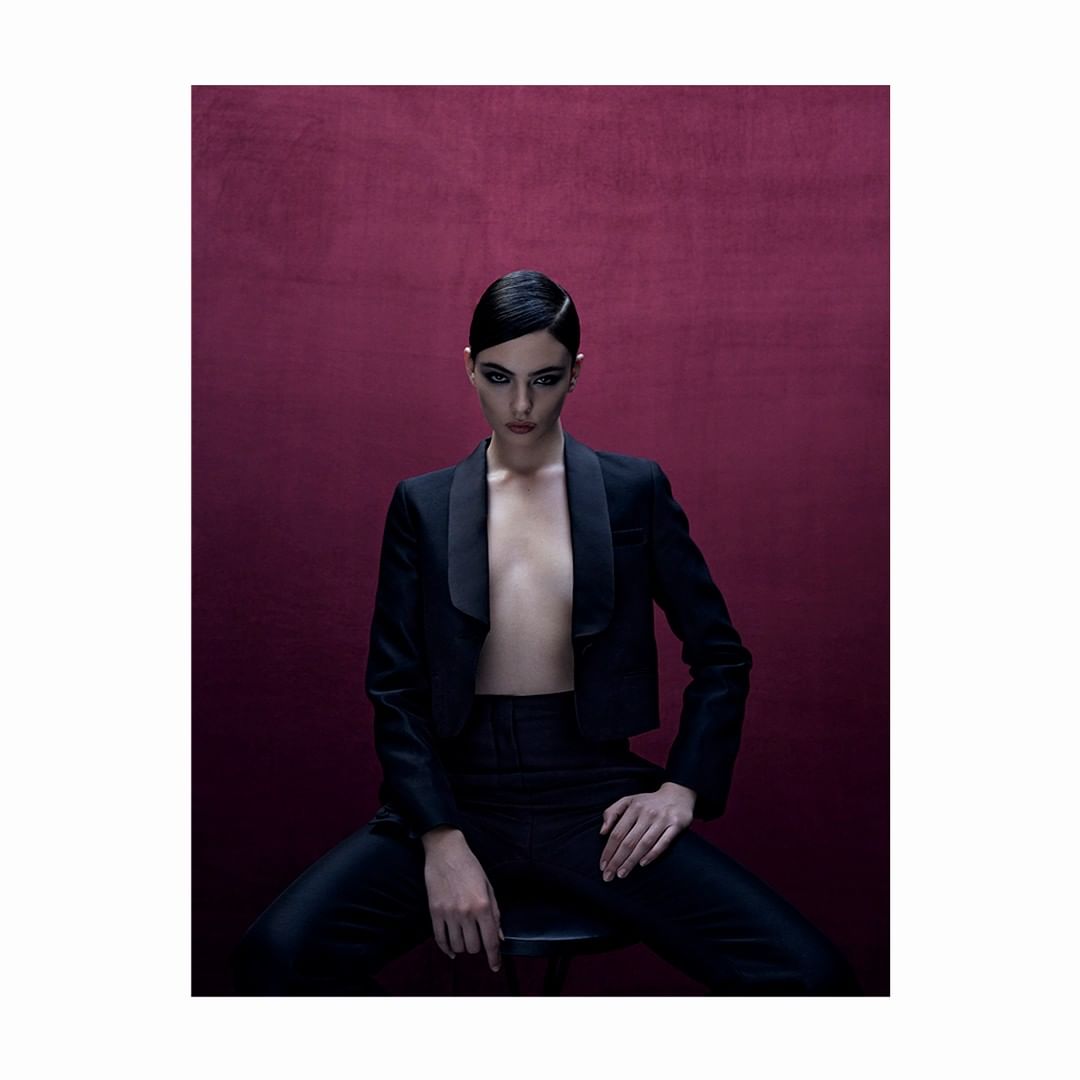 Elegant woman with slicked-back hair poses in a stylish black tuxedo and pants against a deep burgundy background, studio lighting.
