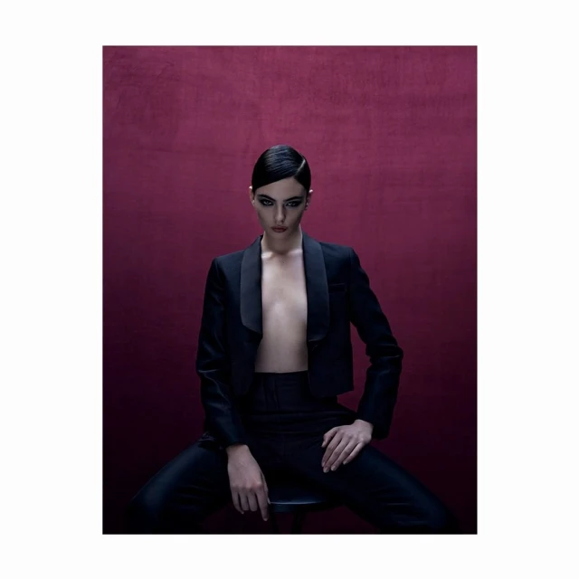 Elegant woman with slicked-back hair poses in a stylish black tuxedo and pants against a deep burgundy background, studio lighting.