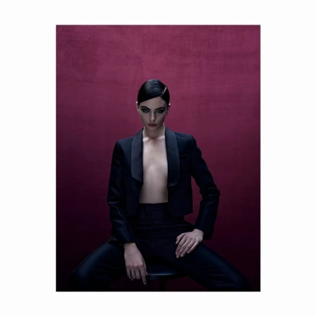 Elegant woman with slicked-back hair poses in a stylish black tuxedo and pants against a deep burgundy background, studio lighting.