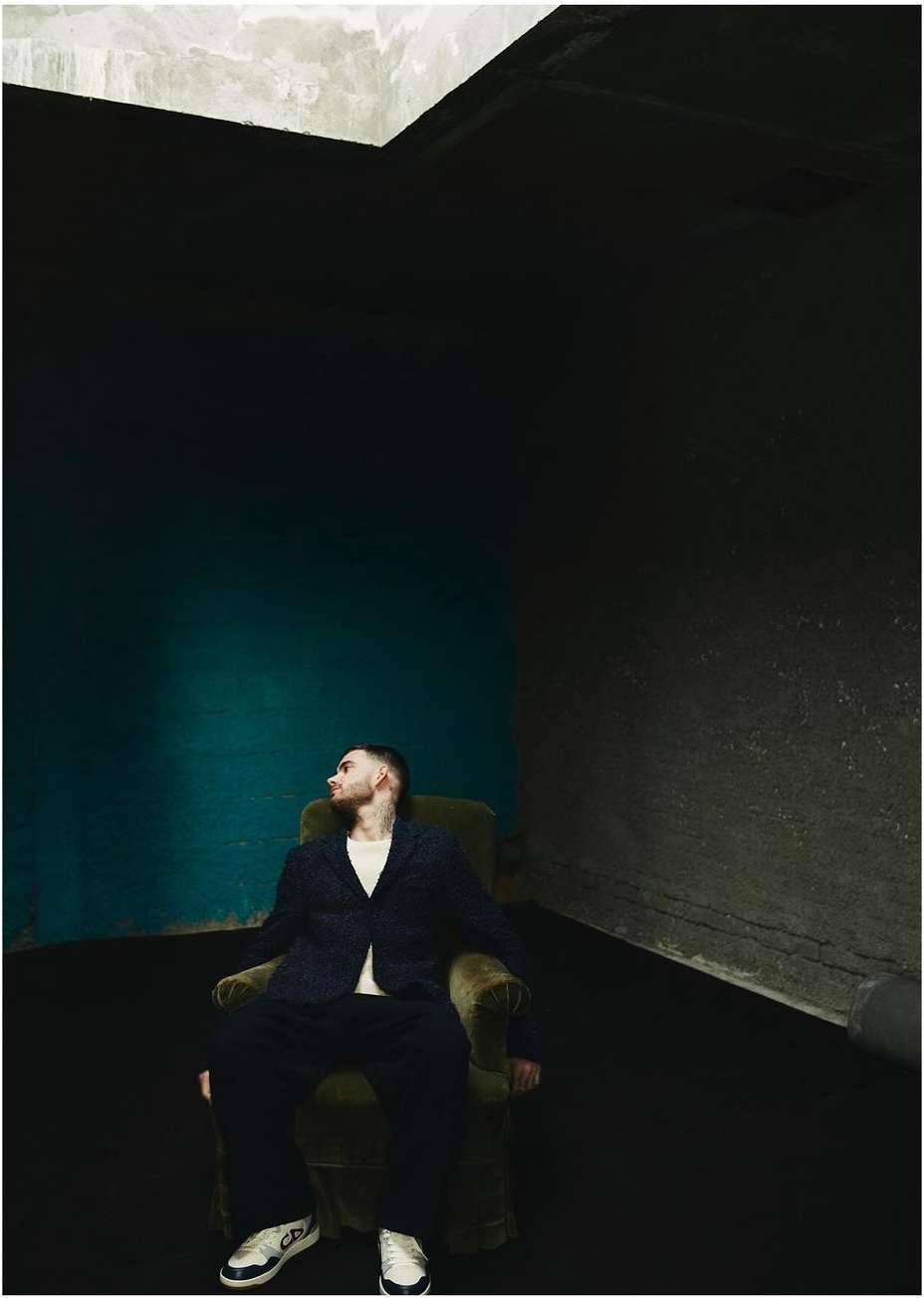 An adult male sits in an olive-green armchair wearing a navy suit jacket, matching trousers, and white sneakers in an indoor studio setting.
