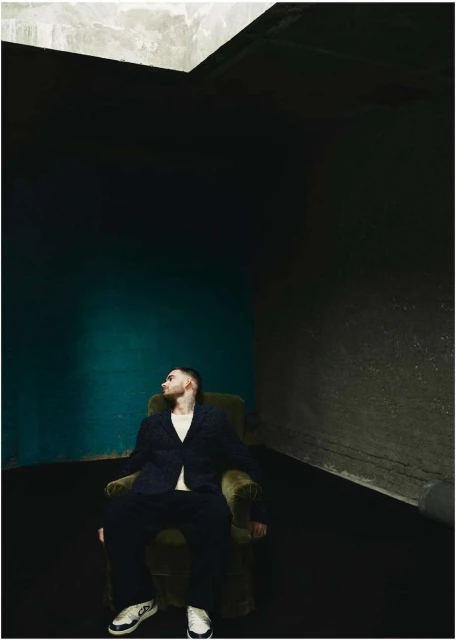 An adult male sits in an olive-green armchair wearing a navy suit jacket, matching trousers, and white sneakers in an indoor studio setting.