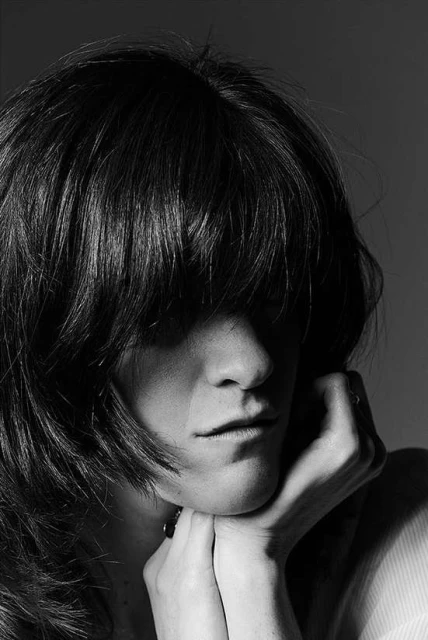 Black and white portrait of a person with long dark hair covering their face, resting their chin on their hands in an indoor studio setting.