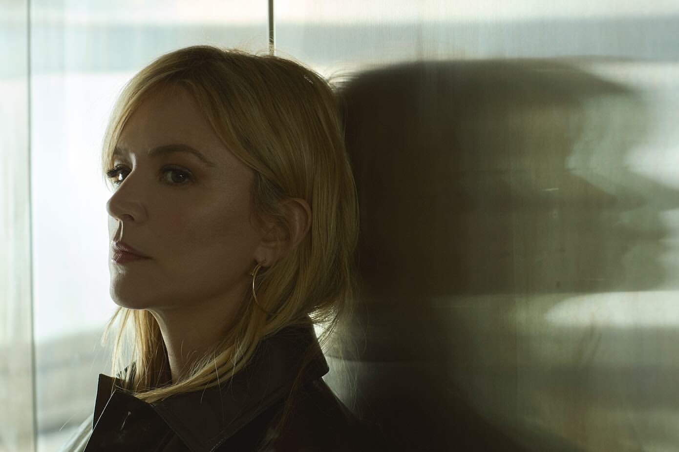 Portrait of a blonde woman in a dark leather jacket leaning against a textured glass wall with a distinct shadow silhouette.