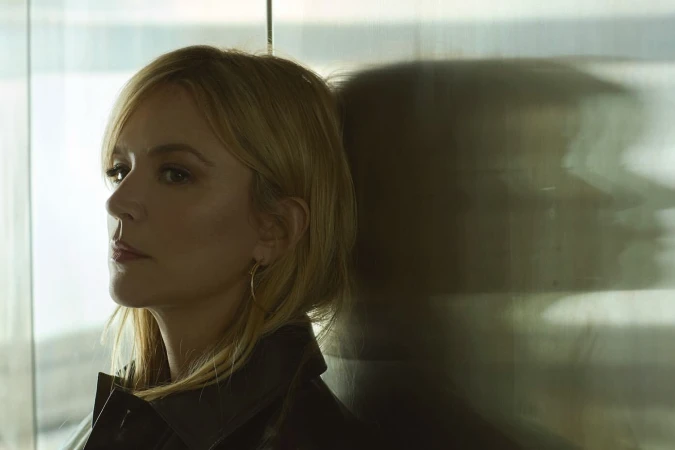 Portrait of a blonde woman in a dark leather jacket leaning against a textured glass wall with a distinct shadow silhouette.
