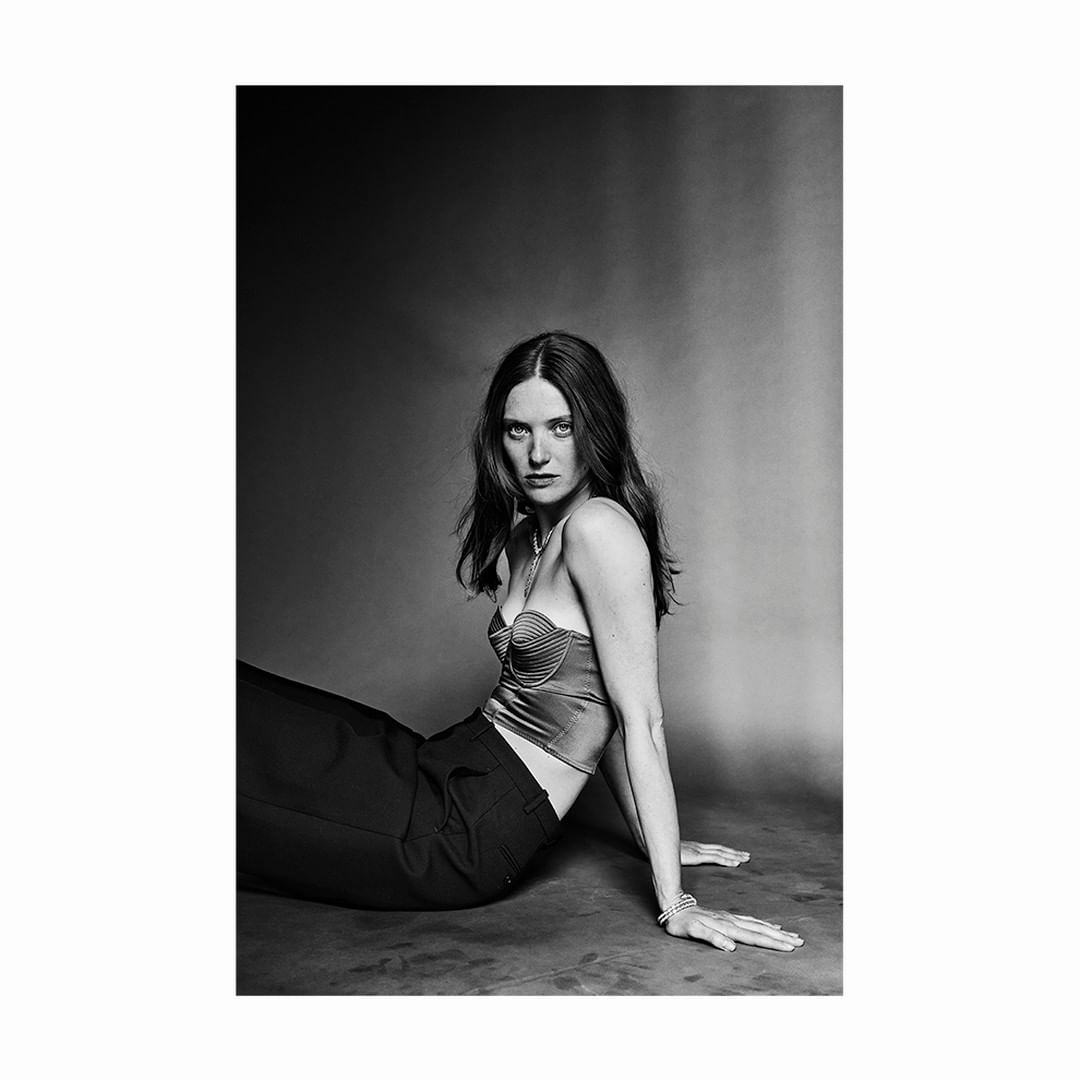 Monochrome portrait of an adult woman sitting on the floor, wearing a bustier top and pants, with an expressive gaze and soft studio lighting.