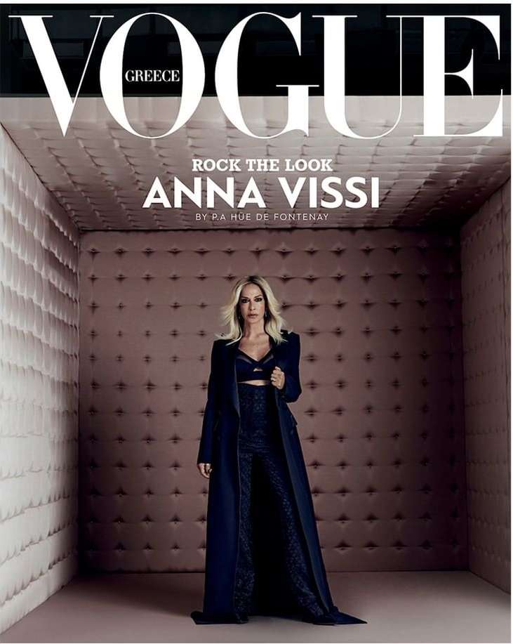 Vogue Greece cover featuring Anna Vissi in a long, dark coat and pants, posing in a padded room with the magazine's title prominently displayed.