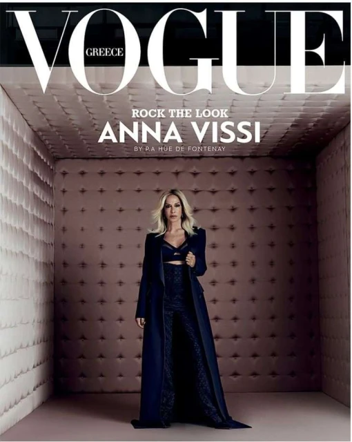 Vogue Greece cover featuring Anna Vissi in a long, dark coat and pants, posing in a padded room with the magazine's title prominently displayed.