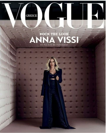 Vogue Greece cover featuring Anna Vissi in a long, dark coat and pants, posing in a padded room with the magazine's title prominently displayed.