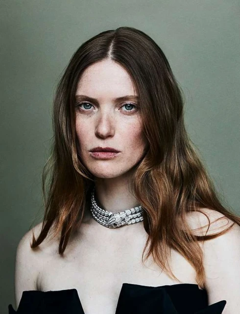 Portrait of a woman with red hair wearing a diamond necklace and a black strapless dress, against a neutral green background.