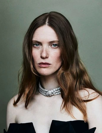 Portrait of a woman with red hair wearing a diamond necklace and a black strapless dress, against a neutral green background.