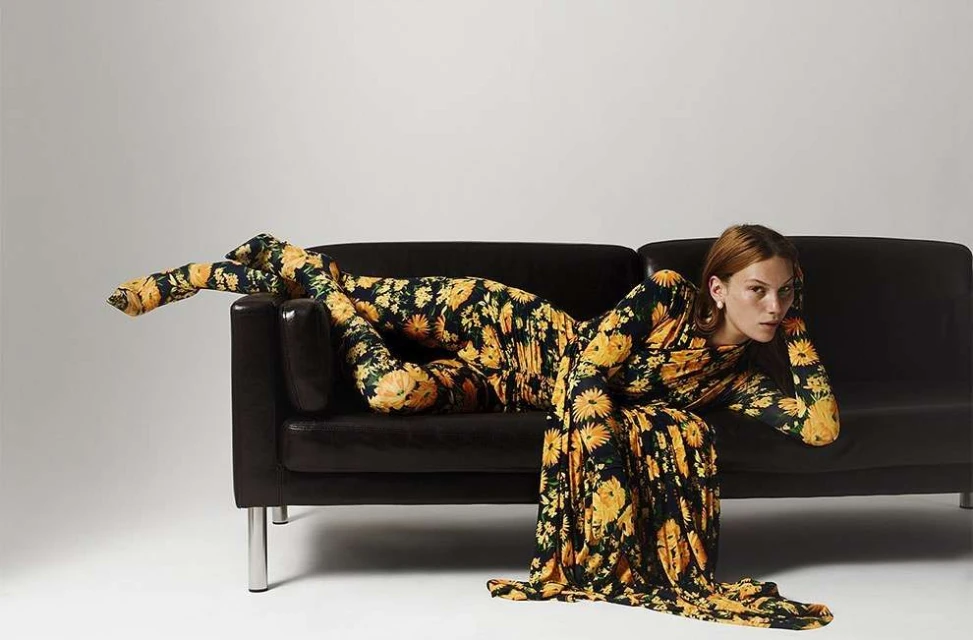 A woman in a floral jumpsuit and dress lies on a black couch in a studio setting, posing for a fashion-style portrait shot.
