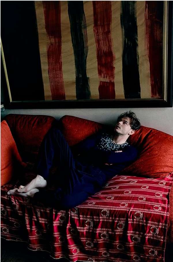 Young man reclines on a red sofa, gazing upward at an abstract painting with red and black stripes, in a softly lit indoor setting.
