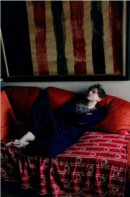 Young man reclines on a red sofa, gazing upward at an abstract painting with red and black stripes, in a softly lit indoor setting.