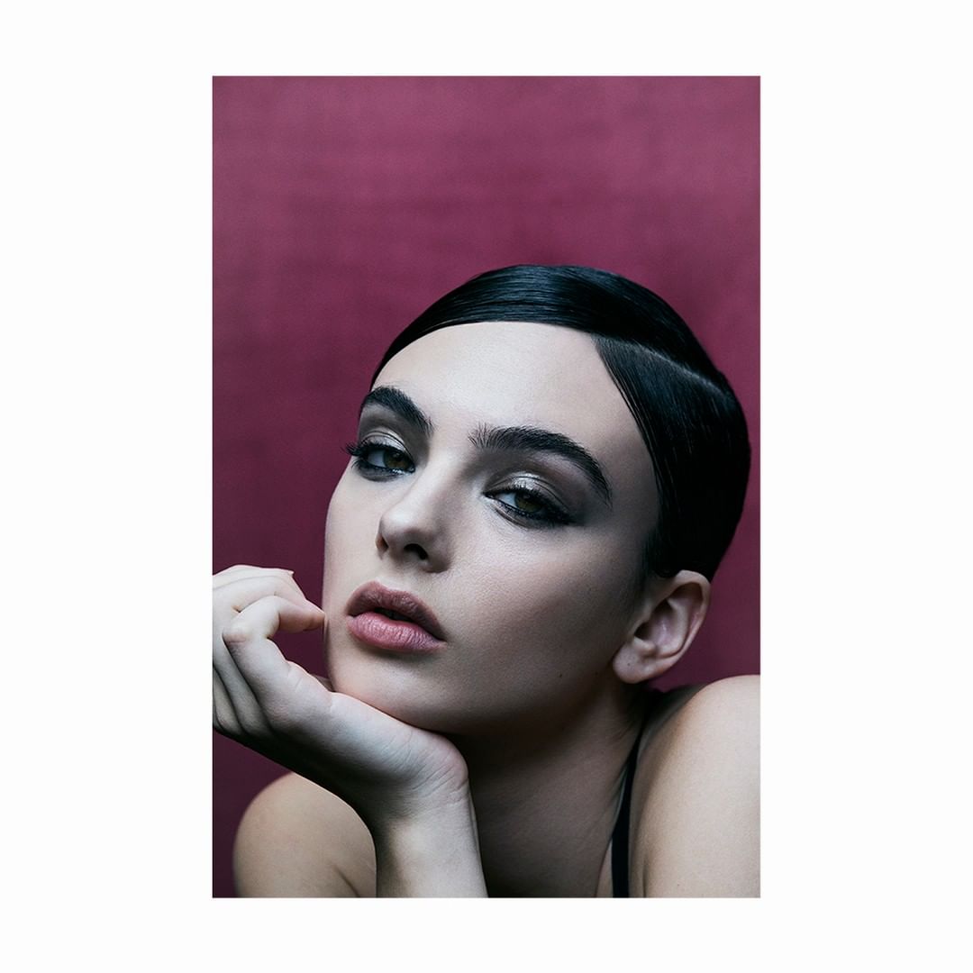 Close-up portrait of a woman with sleek black hair and dramatic eye makeup, posing against a dark red background in a studio setting.
