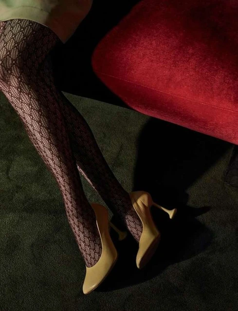 Close-up of tan stiletto heels and patterned black tights on a woman's legs against a dark green textured backdrop and a red cushion.
