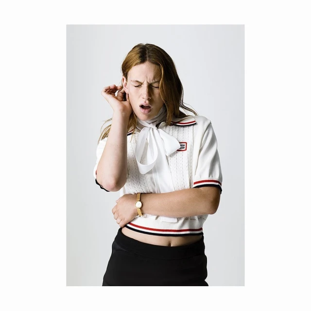 Young woman with red hair in a white top and black skirt is photographed in a studio. She has her fingers in her ear with an open mouth.