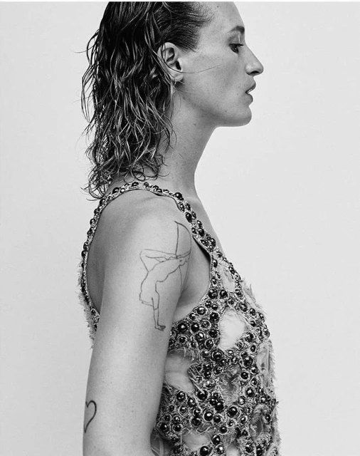 Monochrome portrait of an adult female with wet hair, tattoos, and beaded tank top in a side view, set against a plain background.