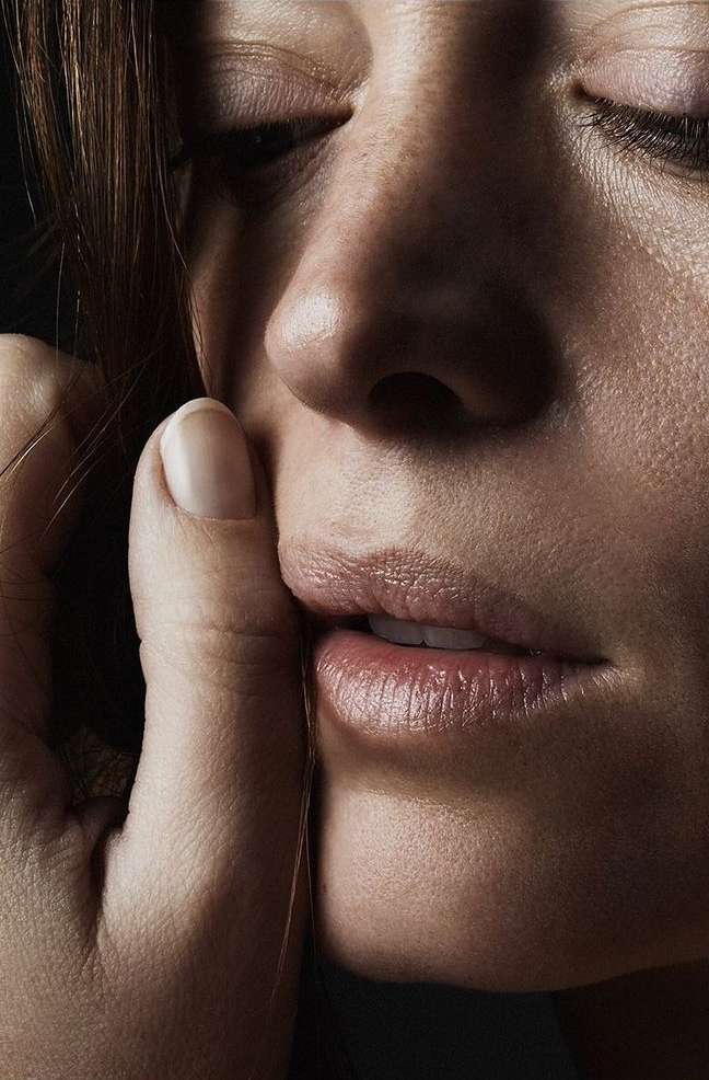 Close-up of a young adult woman's face with her eyes closed and a hand touching her face in a dimly lit indoor setting.
