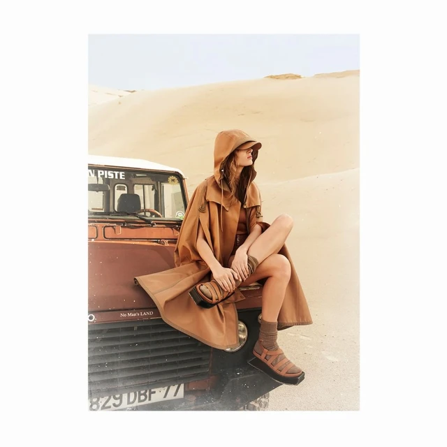 Woman in hooded cape sitting on the hood of a Land Rover in a desert landscape, wearing sandals and socks, fashion photography.