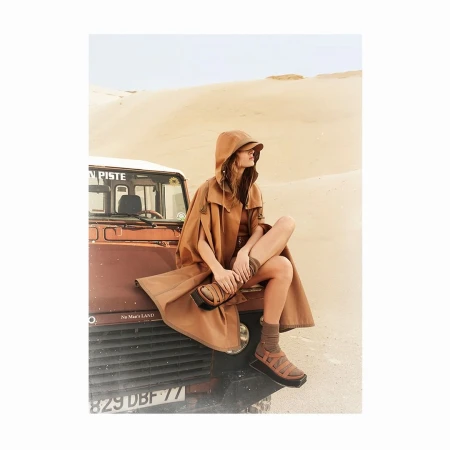 Woman in hooded cape sitting on the hood of a Land Rover in a desert landscape, wearing sandals and socks, fashion photography.