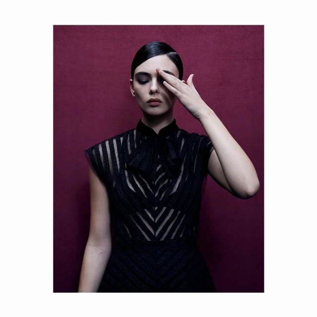 Studio portrait of a woman in a black sheer dress with an elaborate bow, posing with one hand covering her face against a maroon backdrop.