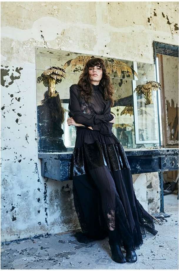 A brunette model in a sheer black dress stands in a derelict room with crumbling walls, ornate mirrors, and dusty surfaces for a fashion shoot.