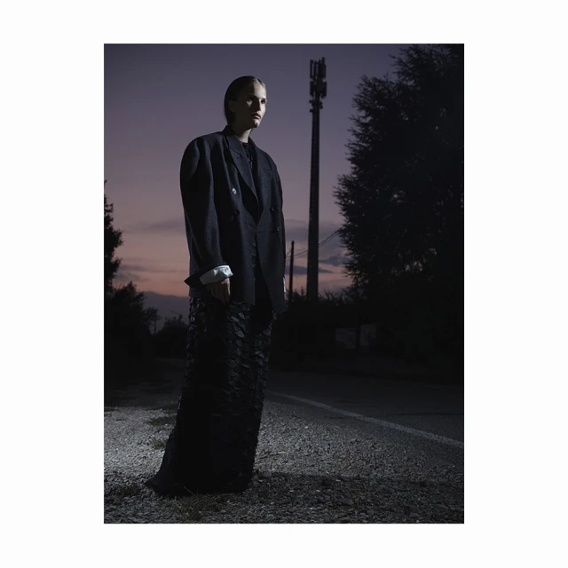 Adult female fashion model wearing a black jacket and black textured dress standing on a road during dusk with communication tower behind.