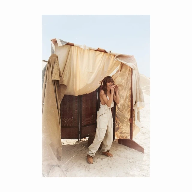 A woman stands in a makeshift shelter in the desert, wearing beige overalls and a white top, smiling in an outdoor fashion shot.