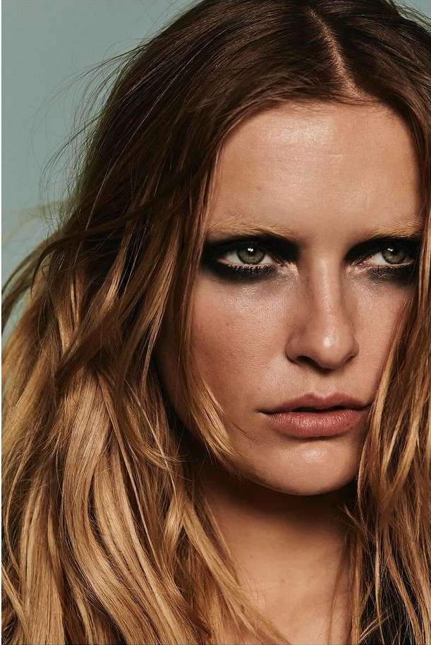 Close-up portrait of a woman with brown hair, heavy dark eye makeup, and neutral lipstick in a studio setting against a light blue backdrop.