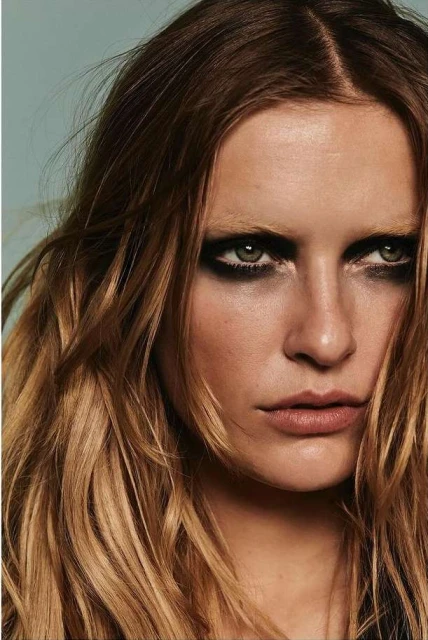 Close-up portrait of a woman with brown hair, heavy dark eye makeup, and neutral lipstick in a studio setting against a light blue backdrop.