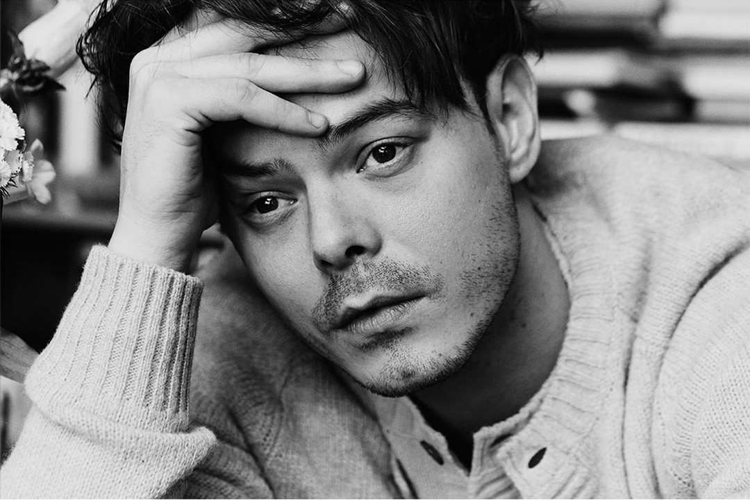 Close-up black and white portrait of a man touching his forehead with his hand while wearing a textured knit sweater