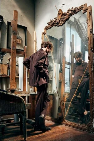 Man in a brown suit stands in an artist's studio, looking at his reflection in an ornate mirror, next to an easel and a wicker chair.