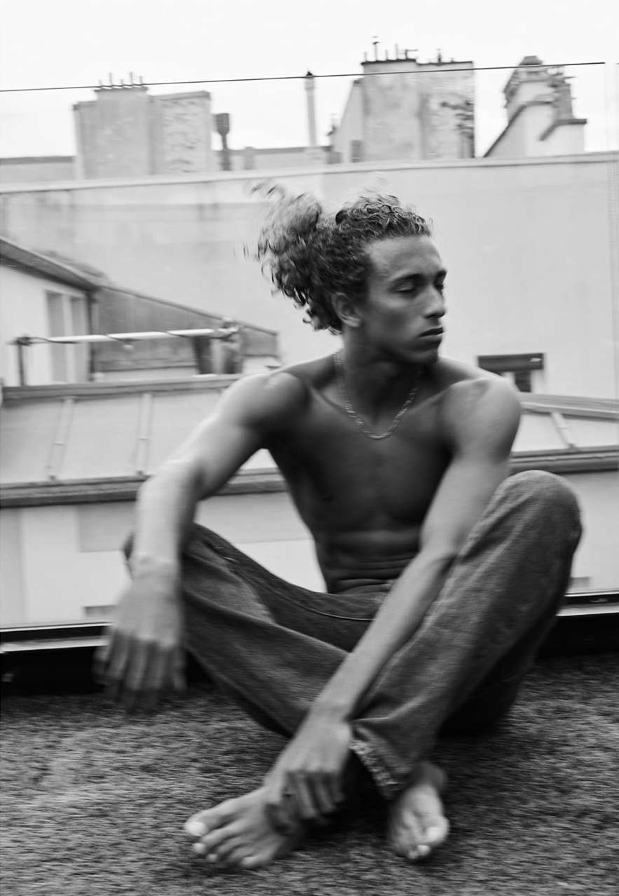 Black and white portrait of a shirtless, barefoot man sitting cross-legged on a rooftop with urban buildings visible in the background.