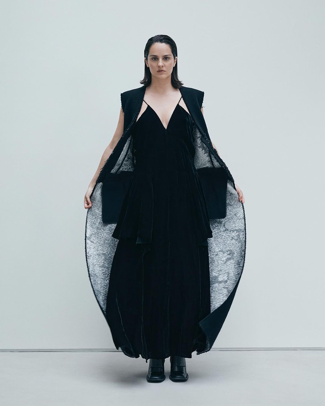 Model wearing a black velvet dress and sleeveless coat against a plain white studio background.
