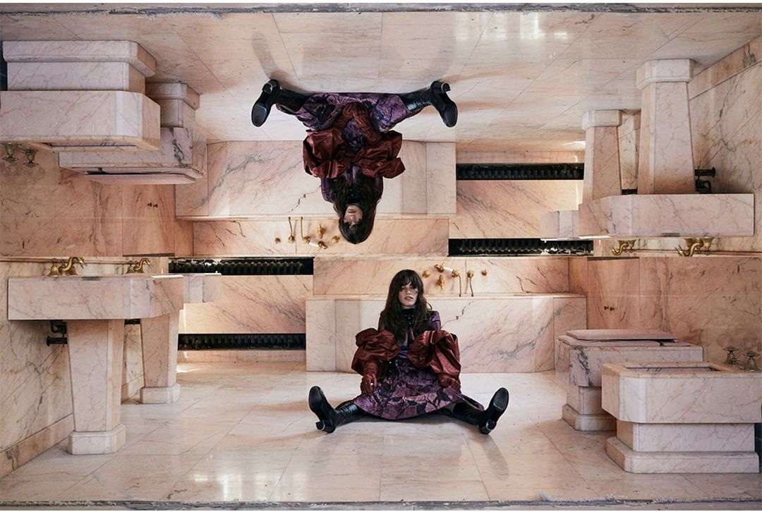 Adult female posing in a marble bathroom setting with reflection, wearing a floral dress, maroon top, and black boots.