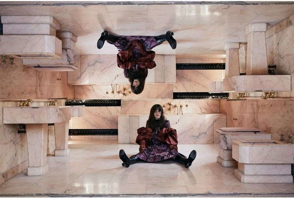 Adult female posing in a marble bathroom setting with reflection, wearing a floral dress, maroon top, and black boots.