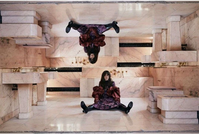 Adult female posing in a marble bathroom setting with reflection, wearing a floral dress, maroon top, and black boots.