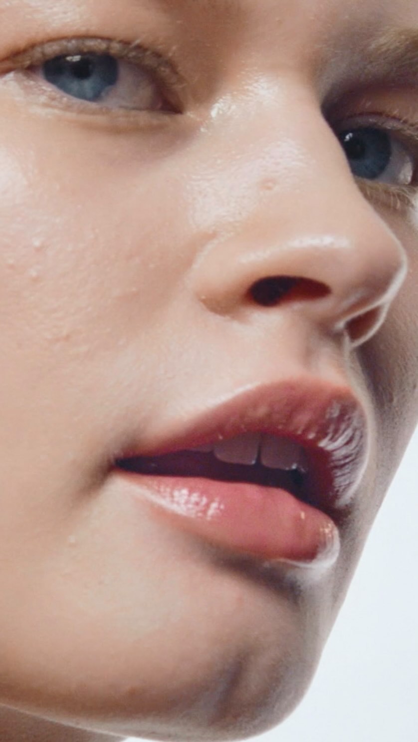 Close-up portrait of a young woman with fair skin, blue eyes, and glossy lips against a bright background, showcasing natural beauty.