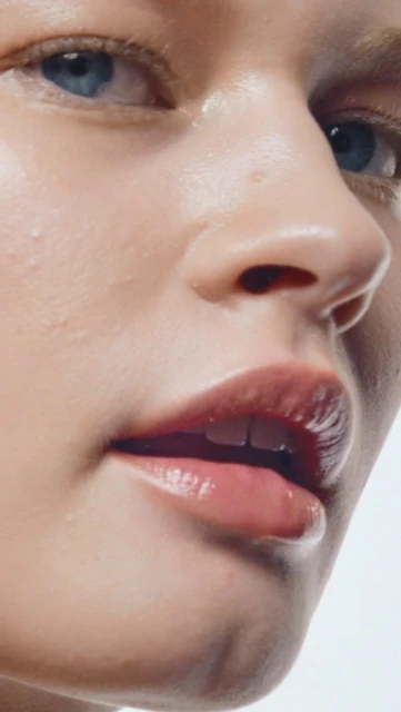 Close-up portrait of a young woman with fair skin, blue eyes, and glossy lips against a bright background, showcasing natural beauty.