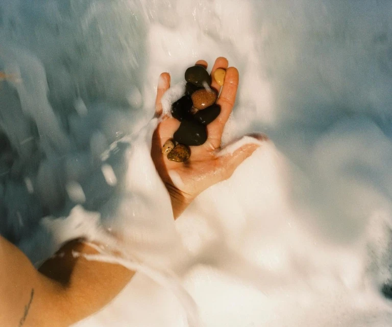 A person's hand is cupped, holding small stones, with foamy, bubbly water surrounding their arm and the rocks.