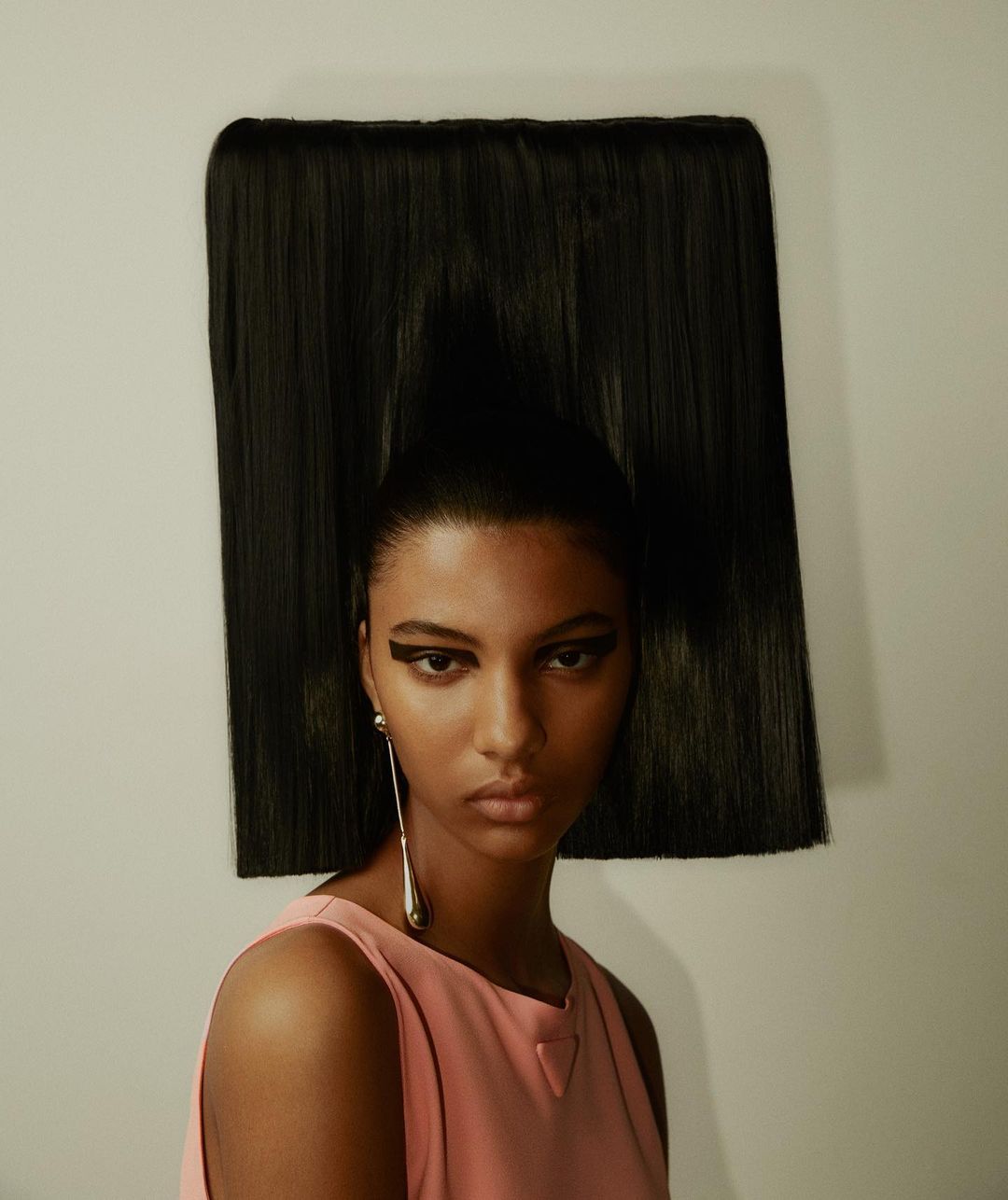 Headshot of a woman with dramatic black eyeliner wearing a pink sleeveless top and a large, rectangular, dark-hair headpiece.